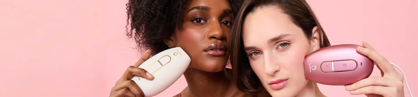 Two women holding RoseSkinCo at-home hair removal devices to their faces against a soft pink background.
