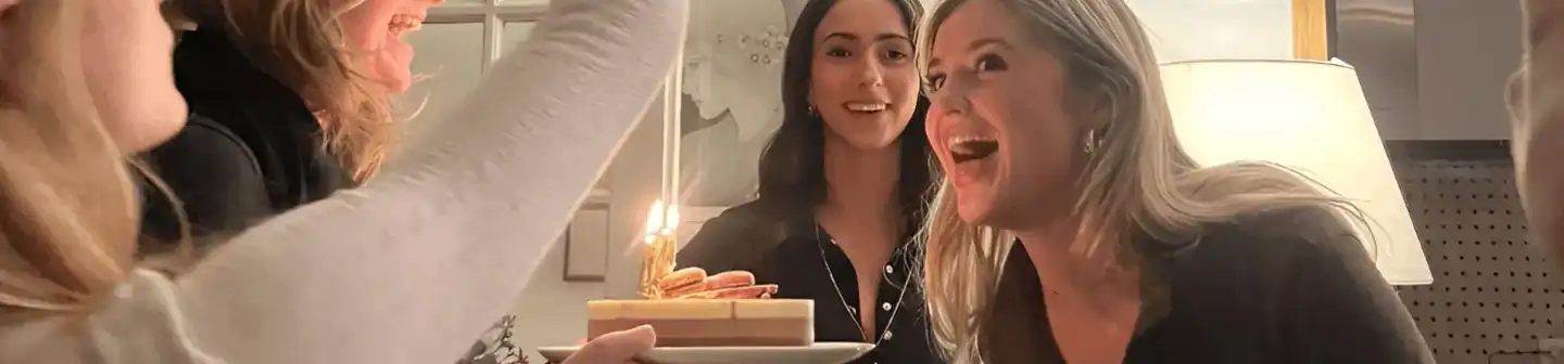 A group of women celebrating together, with one about to blow out a candle on top of a slice of cake topped with macarons.