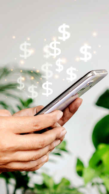 Image of dollar icons rising out of a smart phone being held by two hands in front of some plants.