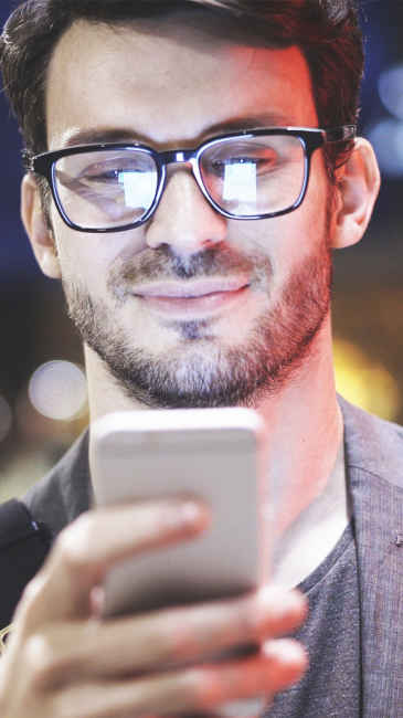 Image of man with his face and glasses illuminated by the smartphone he is holding just below and in front of his face.