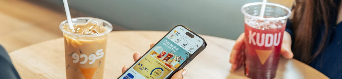 Two people sit at a wooden table, enjoying drinks from KUDU. One woman scrolls through KUDU's rewards app.