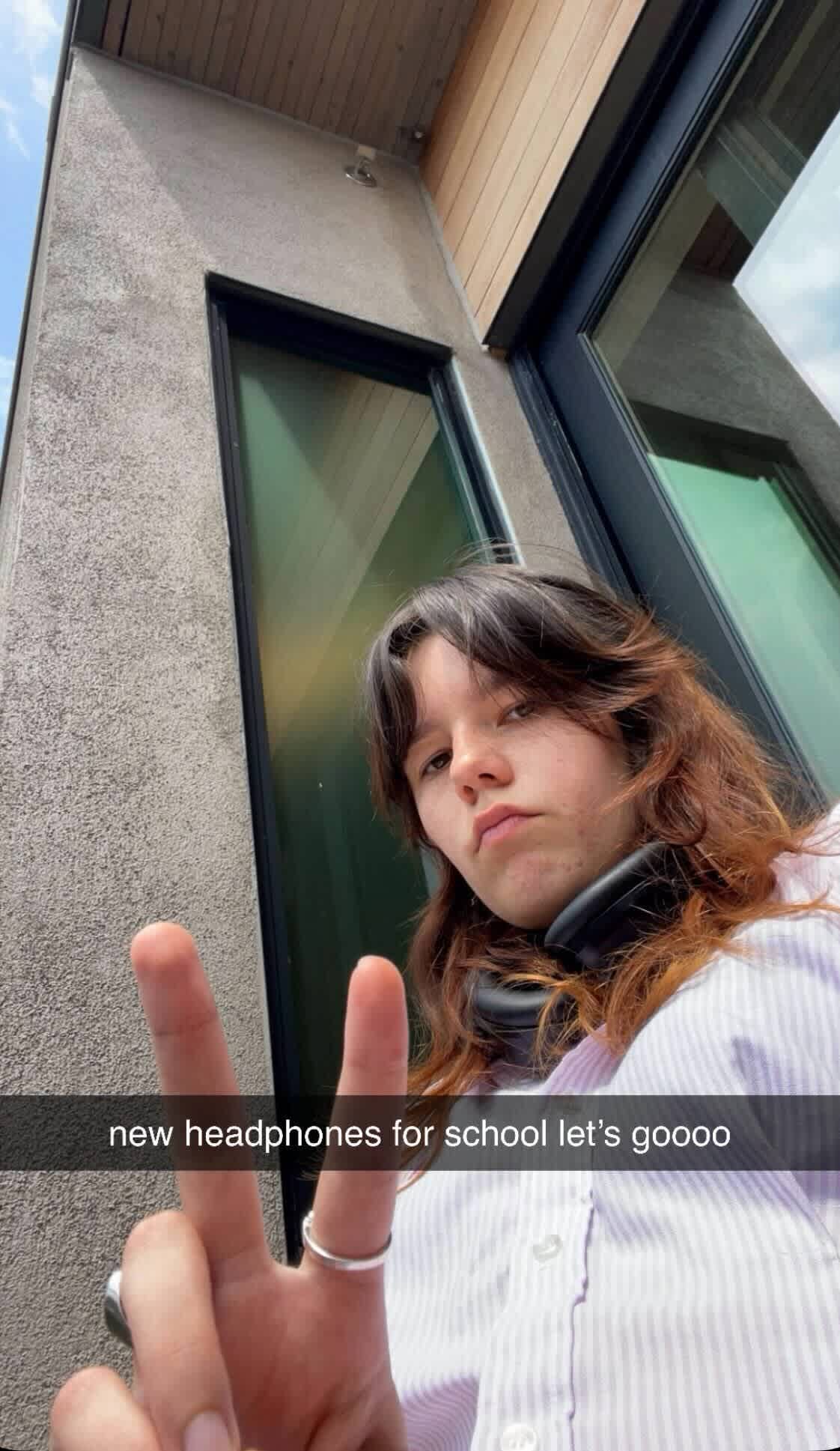 Teen showing new headphones outdoors, sharing excitement with peace sign for social influence on tech purchases.