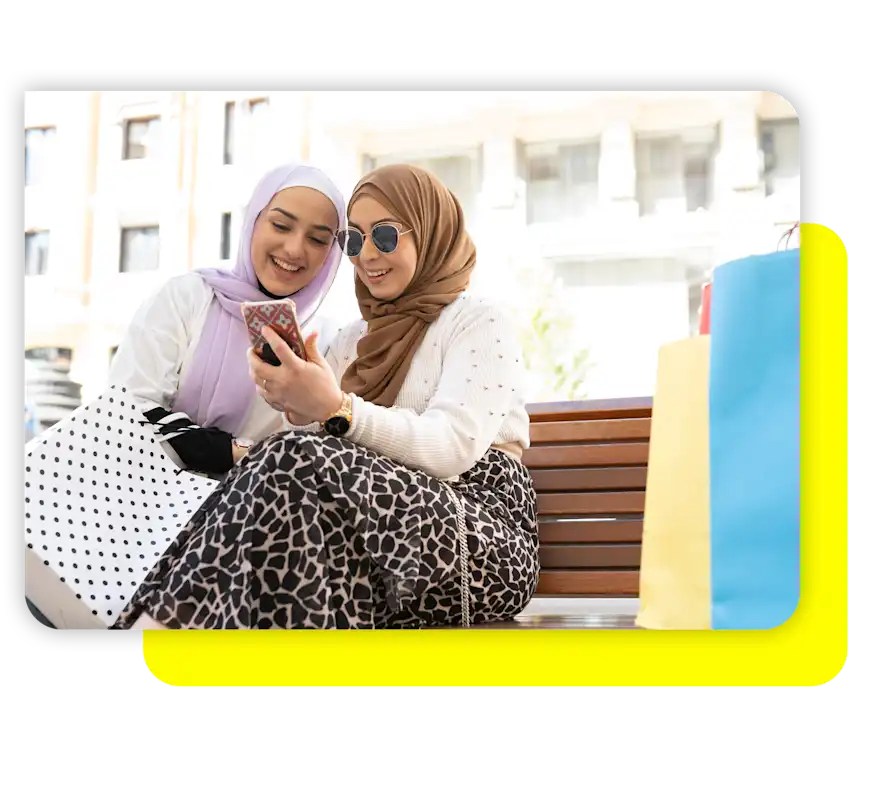 فتاة تتحدث مع صديقتها وتريها الهاتف الجوال بينما تحمل صديقتها حقيبة تسوق.