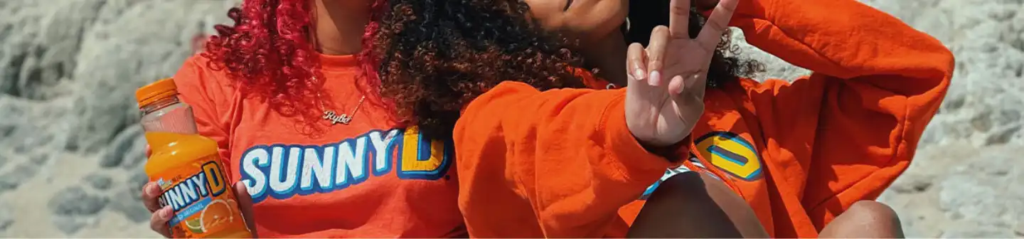 Two women in matching orange SUNNYD shirts pose on a rocky beach, one holding a bottle and the other flashing a peace sign.