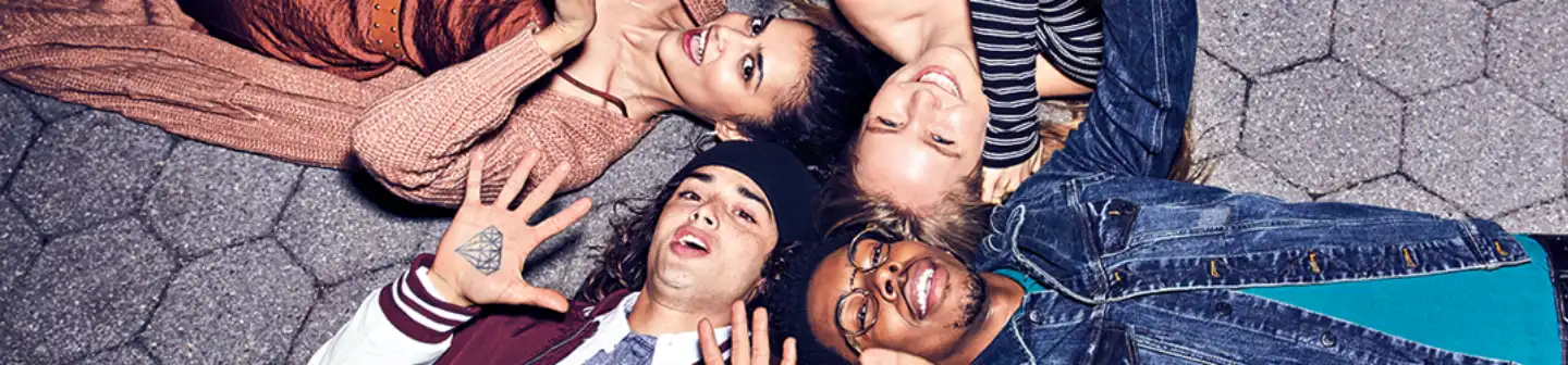 A group of four friends lies on a paved surface, smiling and laughing while dressed in stylish Aéropostale clothing.