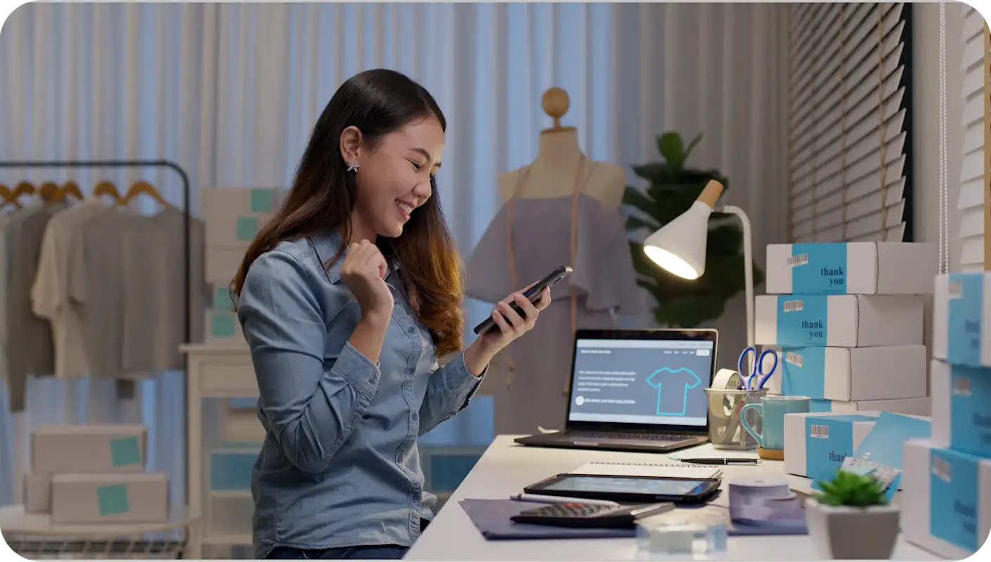 Femme travaillant à un bureau dans un magasin de vêtements, souriant pendant qu'elle regarde son téléphone portable
