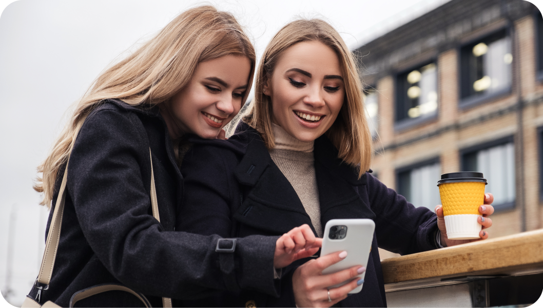 Deux femmes blanches et blondes en manteaux sourient en regardant un téléphone portable dans une rue