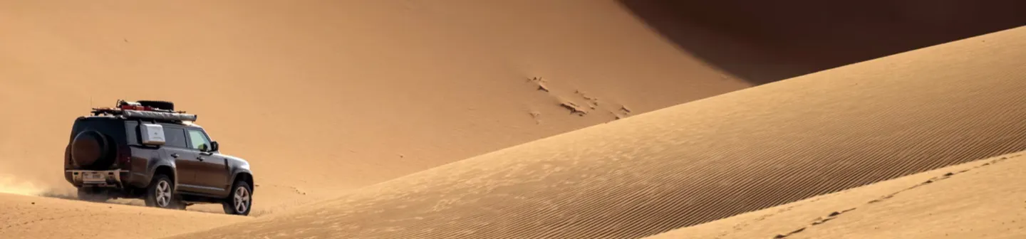 A Defender SUV climbs a steep sand dune, kicking up dust as it traverses a vast, golden desert landscape.
