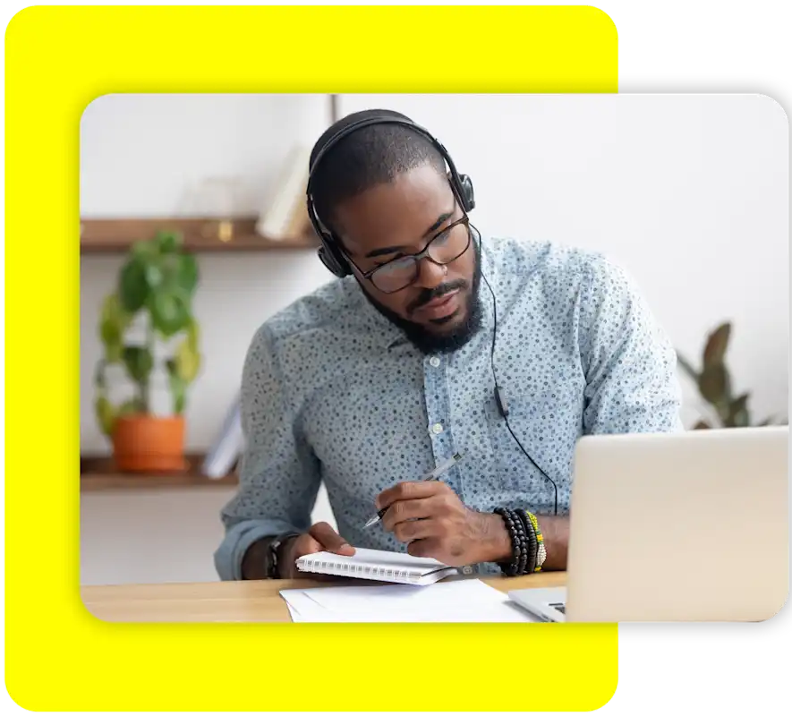 Man with headphones looking at his laptop while writing in a notebook