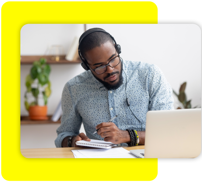 Man with headphones looking at his laptop while writing in a notebook