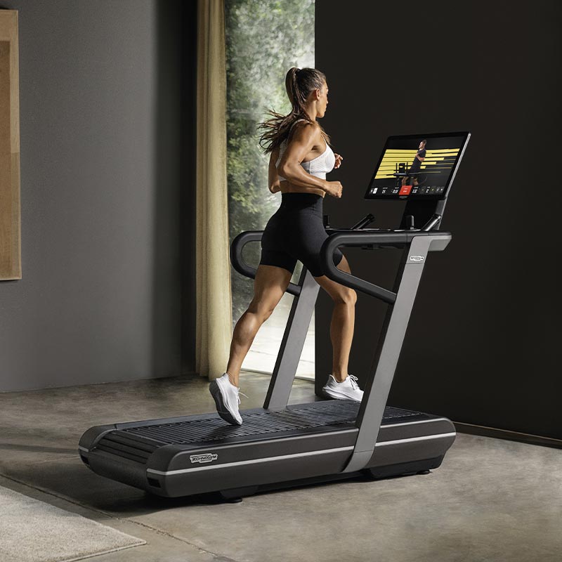 A woman running on a Technogym Run treadmill with a trainer guiding her from the large display
