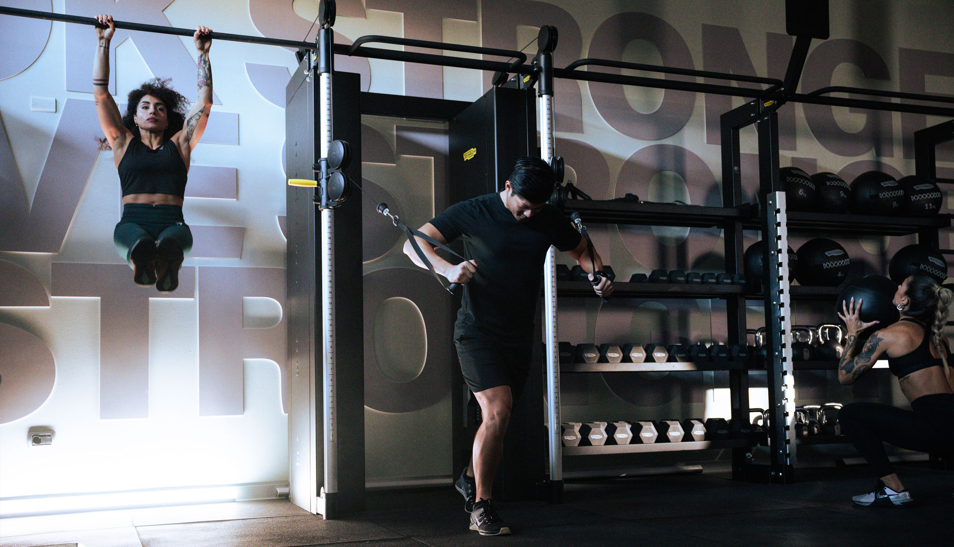 Two young ladies and a young man training on Technogym Universe equipment. The lady on the left is hanging from a bar holding her legs straight, extended at a 90 degrees angle, while the man is about to perform a chest press with the dual adjustable pulley cable machine, and the lady on the right is squatting with a ball in her hand, and about to throw it to the target above her