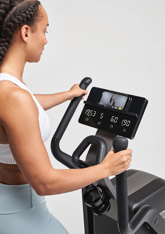 Close-up of a person using a Technogym Elliptical cardio machine with digital console displaying workout metrics and a smartphone streaming a training video