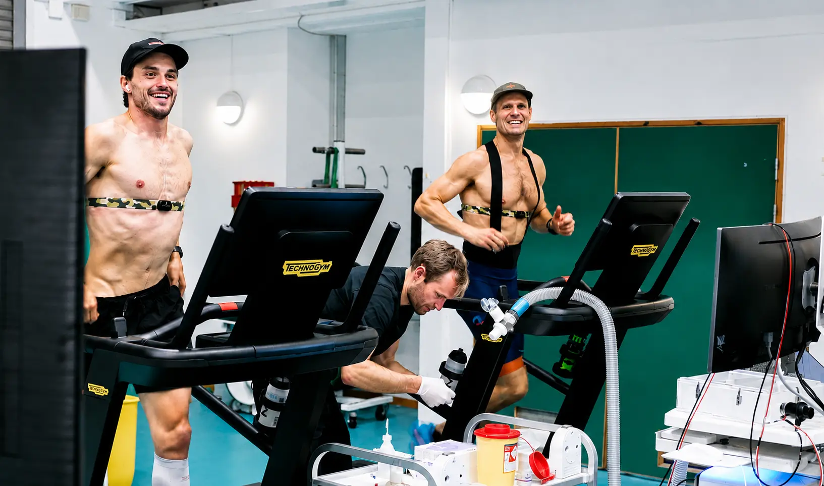 Two athletes undergo performance testing on Technogym Skillrun treadmills, wearing heart rate monitors while a technician tracks fitness data in a lab.