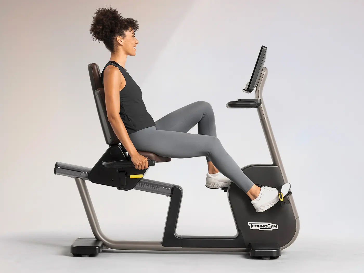 Person demonstrating the walk‑through frame of the Technogym Excite Recline bike, showcasing the low step‑in design for easy access and comfortable positioning during recumbent cycling workouts