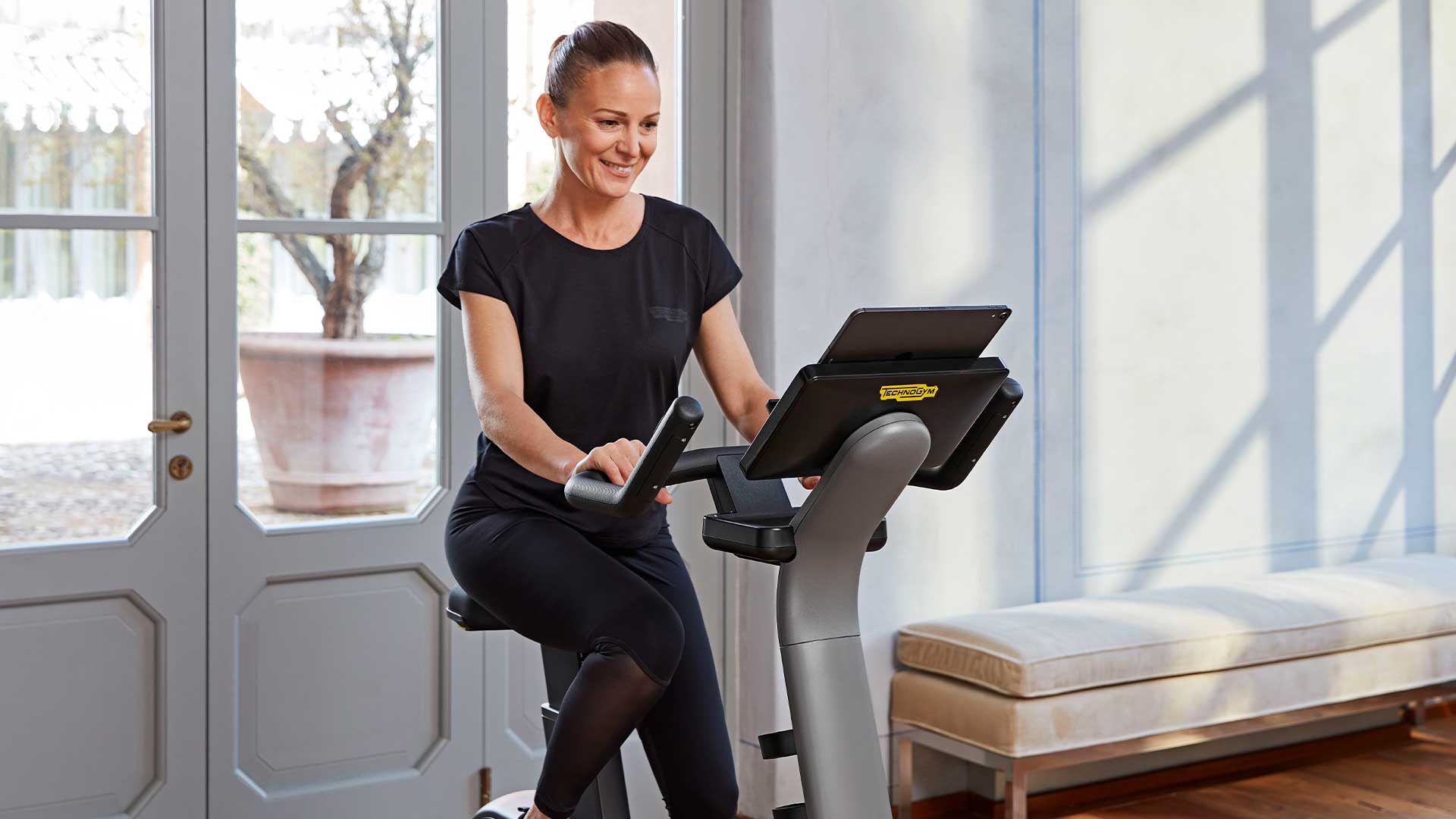 Person using a Technogym exercise bike indoors for a home cardio workout, featuring a sleek design and a dashboard holding a large tablet for interactive training