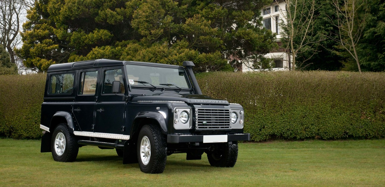 Black Land Rover hearse parked on grass in front of a hedge