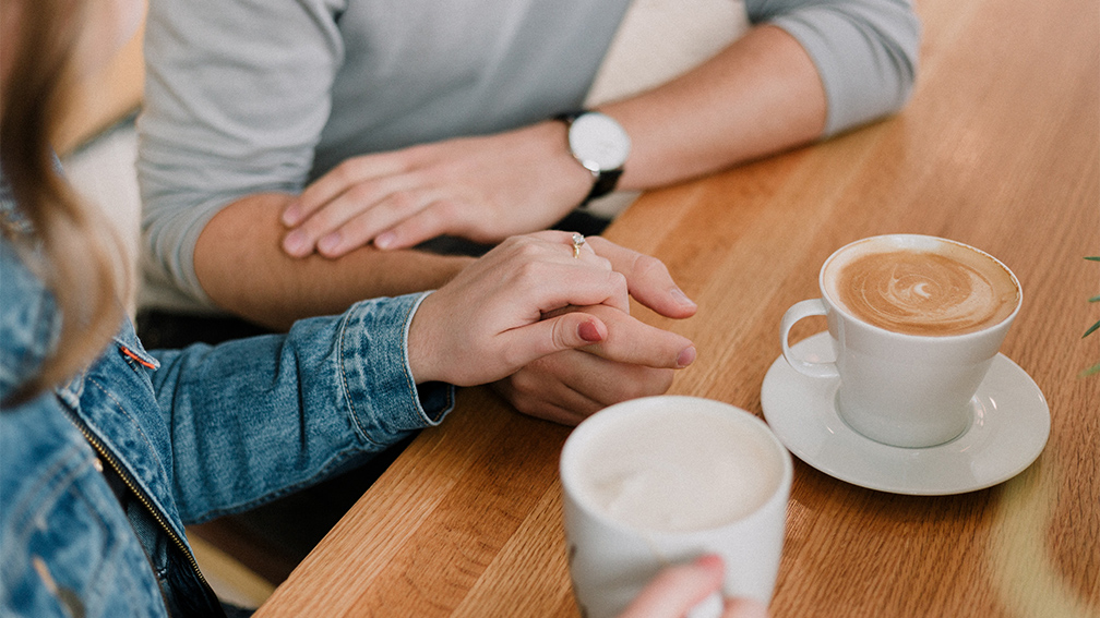 A man and woman sat down, holding hands with a coffee on the table.