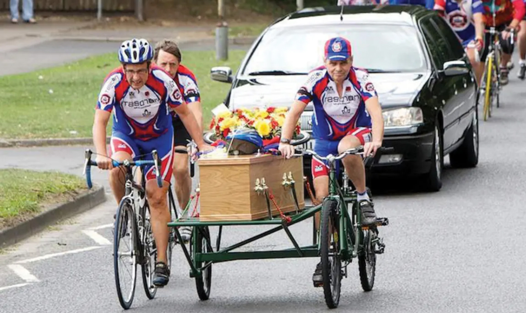 Funeral procession with a coffin with helmet and floral arrangement on a hearse pulled by bicycles 