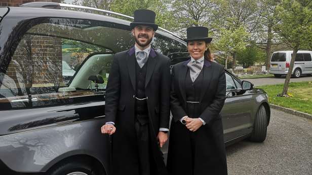 Husband and wife stood in front of a hearse. 