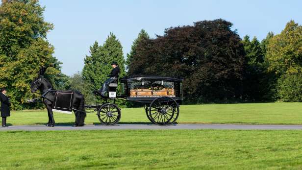 Glass horse drawn hearse with a coffin inside stopped in parkland with a black horse and attendants