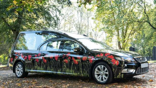 Black hearse with poppies on parked in a woodland with an attendant in front