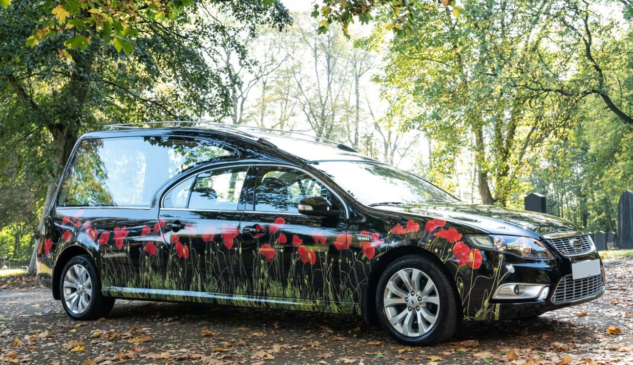 Black hearse with poppies on parked in a woodland with an attendant in front