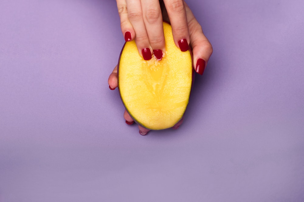 Hand with red finger nails holding half a mango on purple background