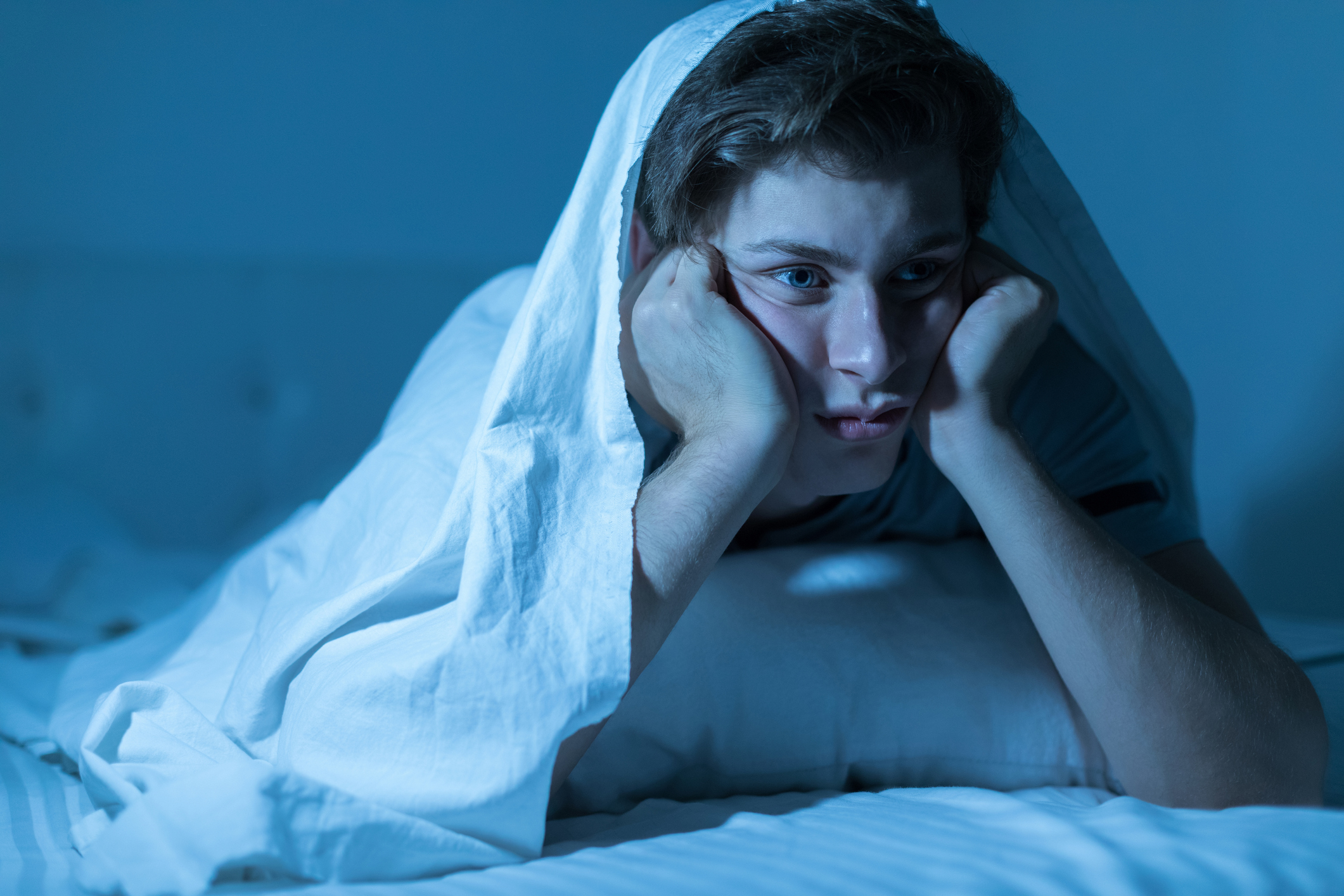 Man with insomnia and trouble sleeping sitting up in bed underneath a sheet