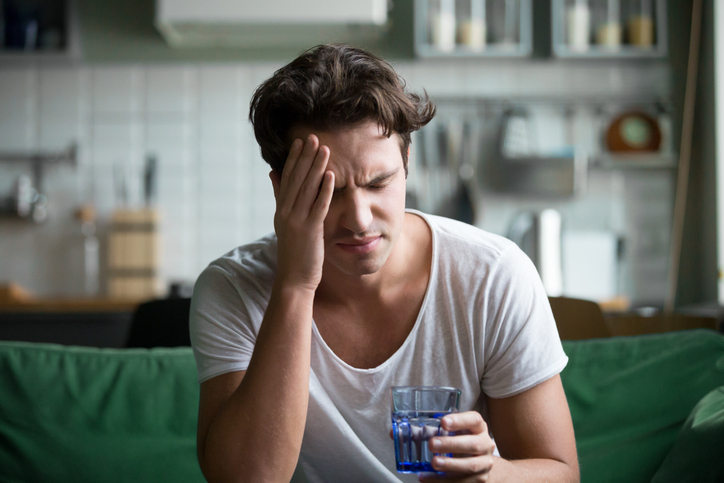 Young man with hangover headache sitting with glass of water in the kitchen