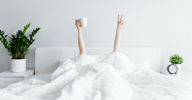 Woman's arms poking out from under duvet giving peace sign holding coffee after great night's sleep