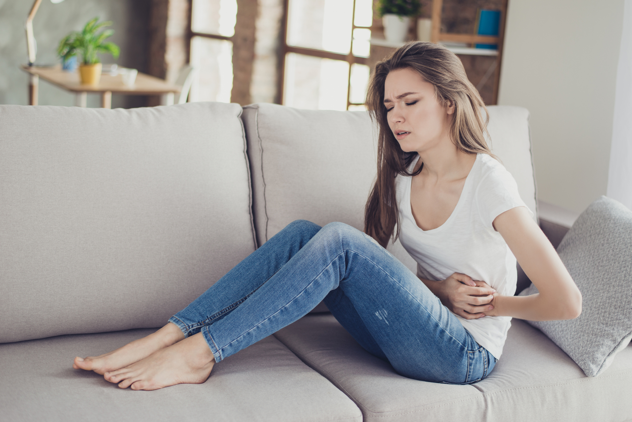 Young woman on a sofa holding her stomach because of pain related to constipation. Credit: Deagreez