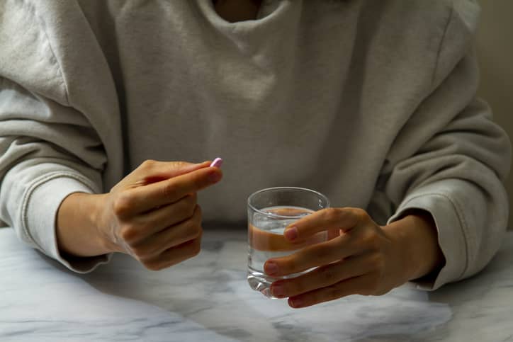 Person in grey sweatshirt holding glass of water and emergency contraceptive pill