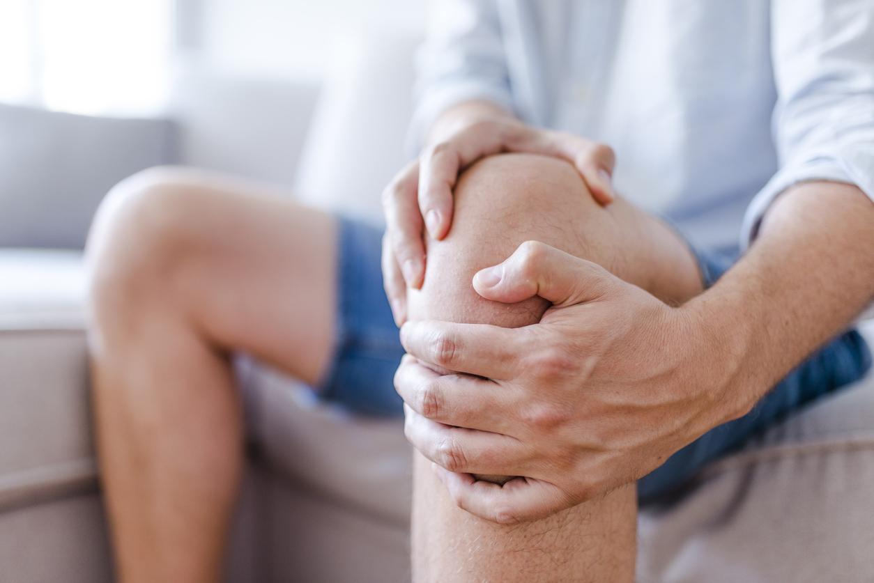 Close-up of man's knee as he sits on a sofa, holding knee in pain