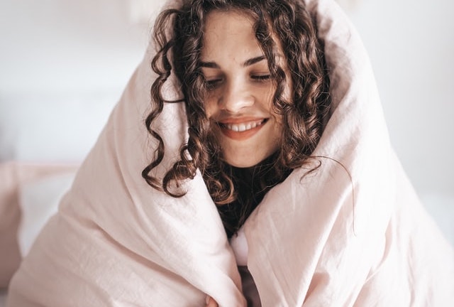 Person wrapped up in a  light pink blanket sitting on their bed