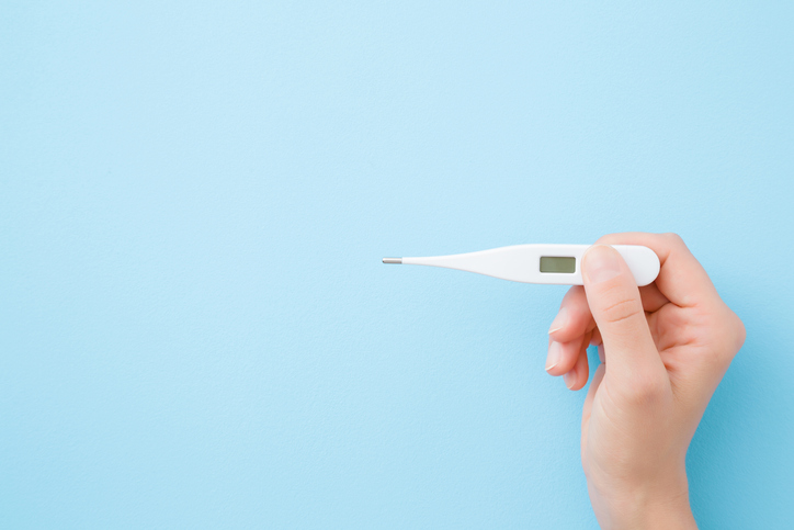 Hand holding white digital thermometer on pastel blue background.