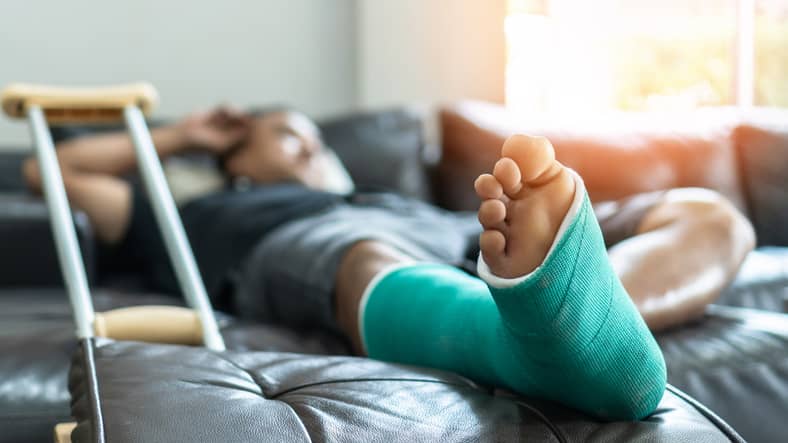 Person lying on sofa with broken leg in a cast beside a crutch