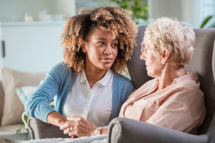 Woman carer talking with elderly woman sitting on armchair in living room