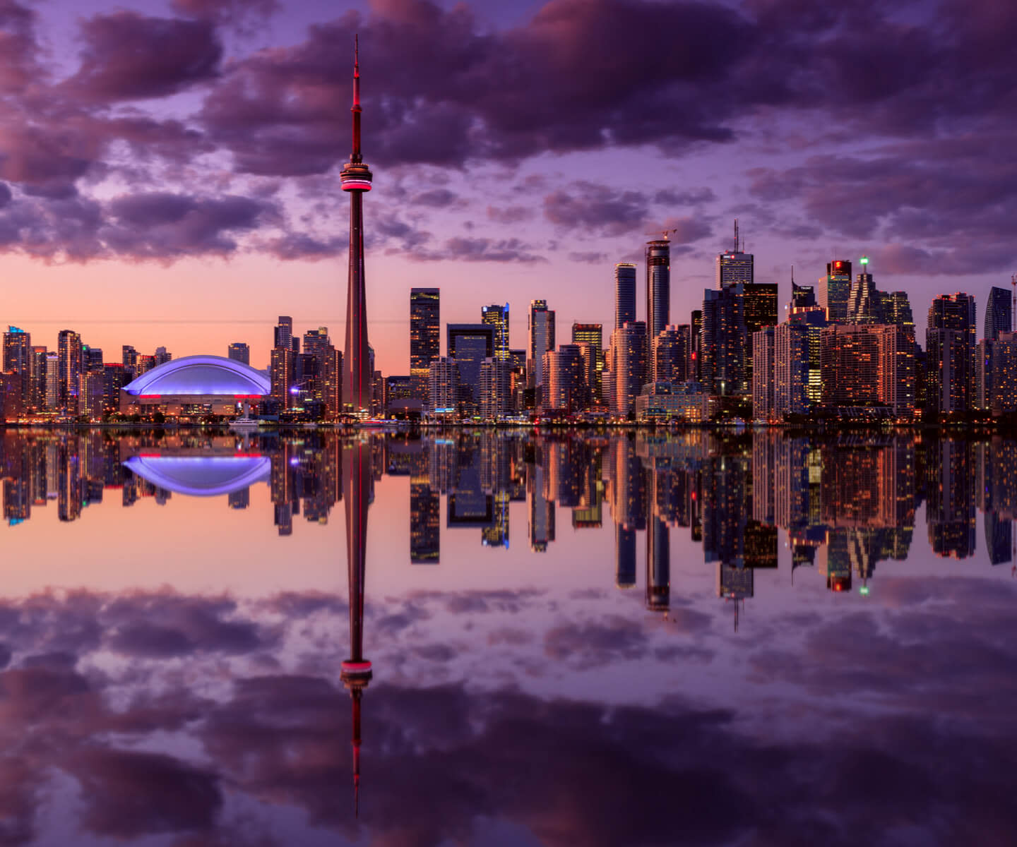 Toronto skyline at dusk, featuring the CN Tower and Rogers Centre, reflects on calm water