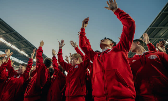 A group of people in red jackets at a stadium enthusiastically raise their arms under a clear sky