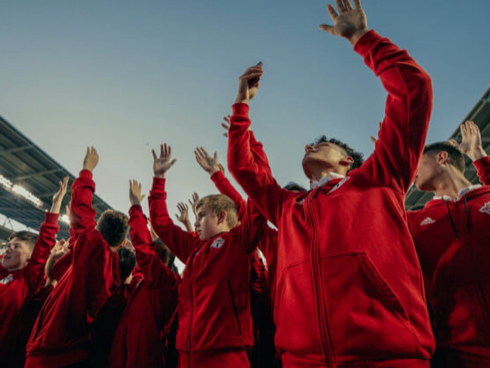 A group of people in red jackets at a stadium enthusiastically raise their arms under a clear sky