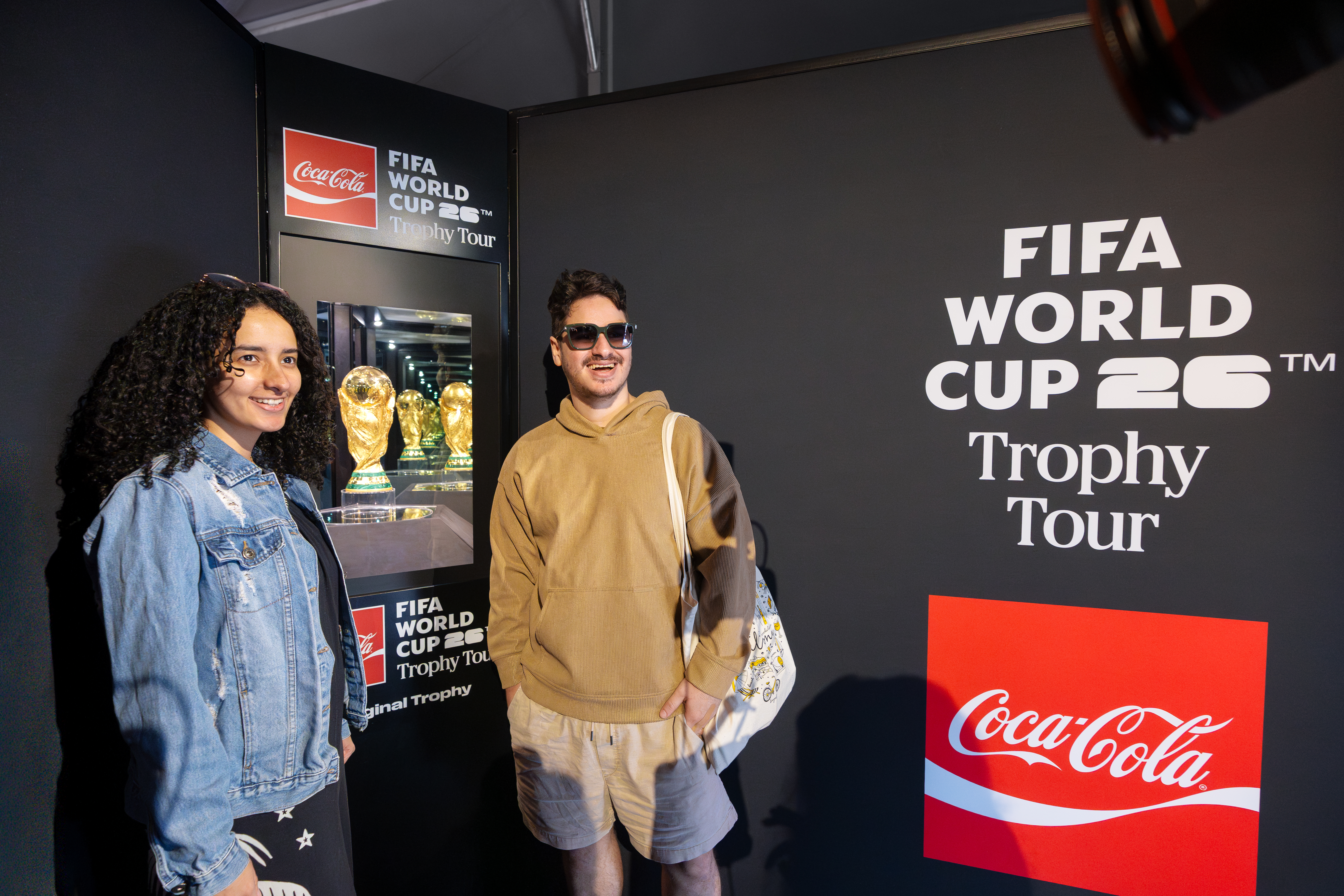 Two people pose for a photo with the FIFA World Cup trophy which sits inside a case surrounded by Coca-Cola branding.