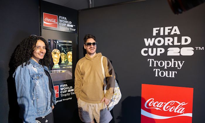 Two people pose for a photo with the FIFA World Cup trophy which sits inside a case surrounded by Coca-Cola branding.