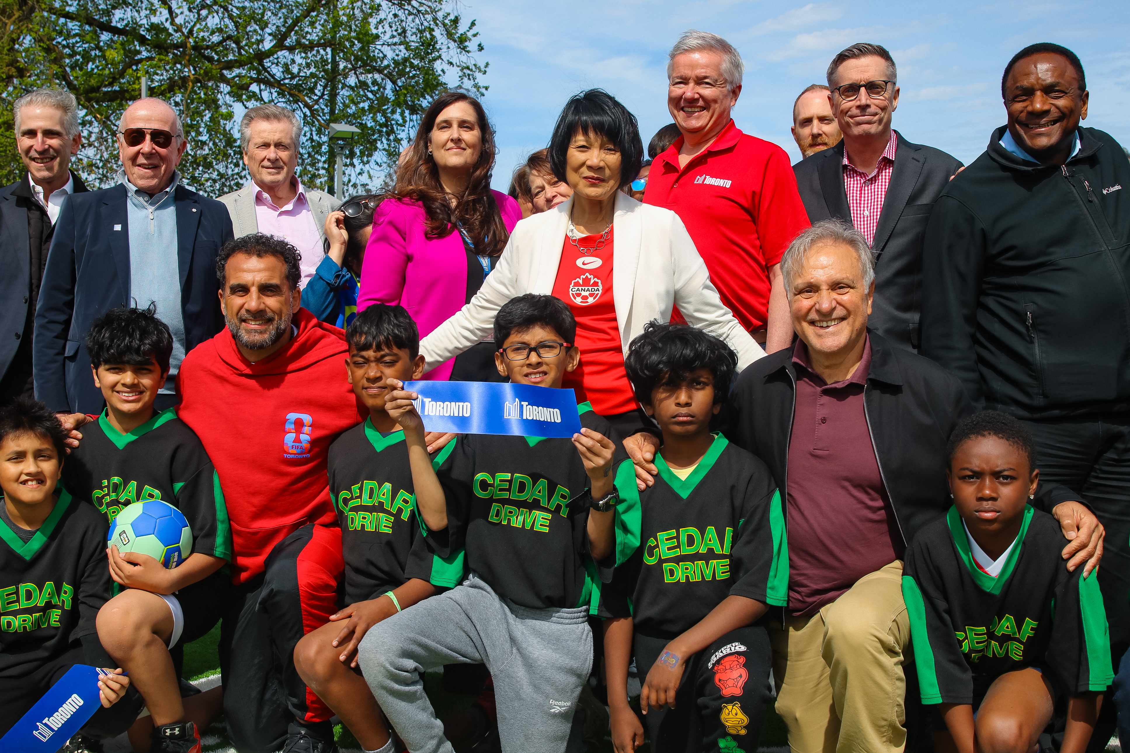 A large group of adults and kids soccer team posing for a photo. A kid in the middle holds up a piece of a cut blue ribbon that reads "Toronto"