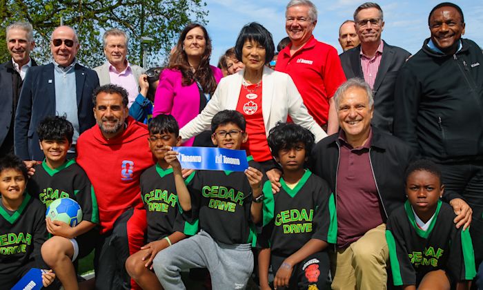 A large group of adults and kids soccer team posing for a photo. A kid in the middle holds up a piece of a cut blue ribbon that reads "Toronto"