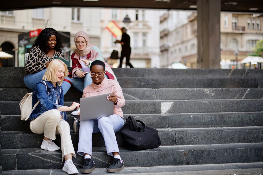 Four students sit on the steps of a university campus.  One has a laptop and is showing something to the other students via the screen.