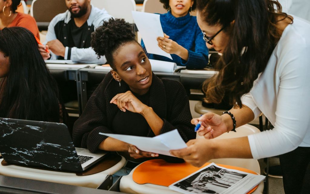 In a busy classroom a student is getting some help from an instructor, the student is listening and being attentive as the instructor explains while referencing a sheet of paper.