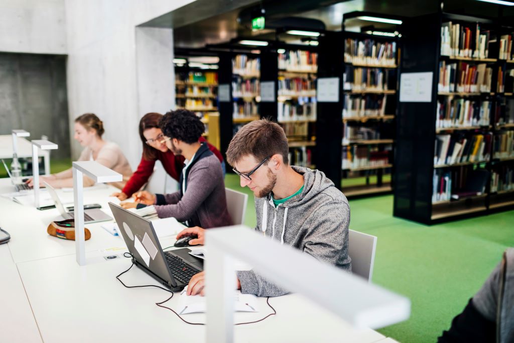 A library with bookshelves in the background, several students sit at a table in the foreground.  One is being helped by a librarian.