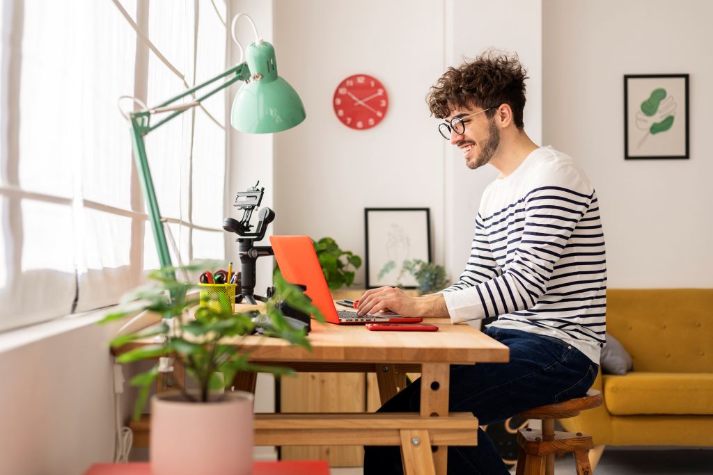 a rustic desk in a contemporary room with an sofa, plant and desk lamp.  A student sits at the desk working on an orange laptop.