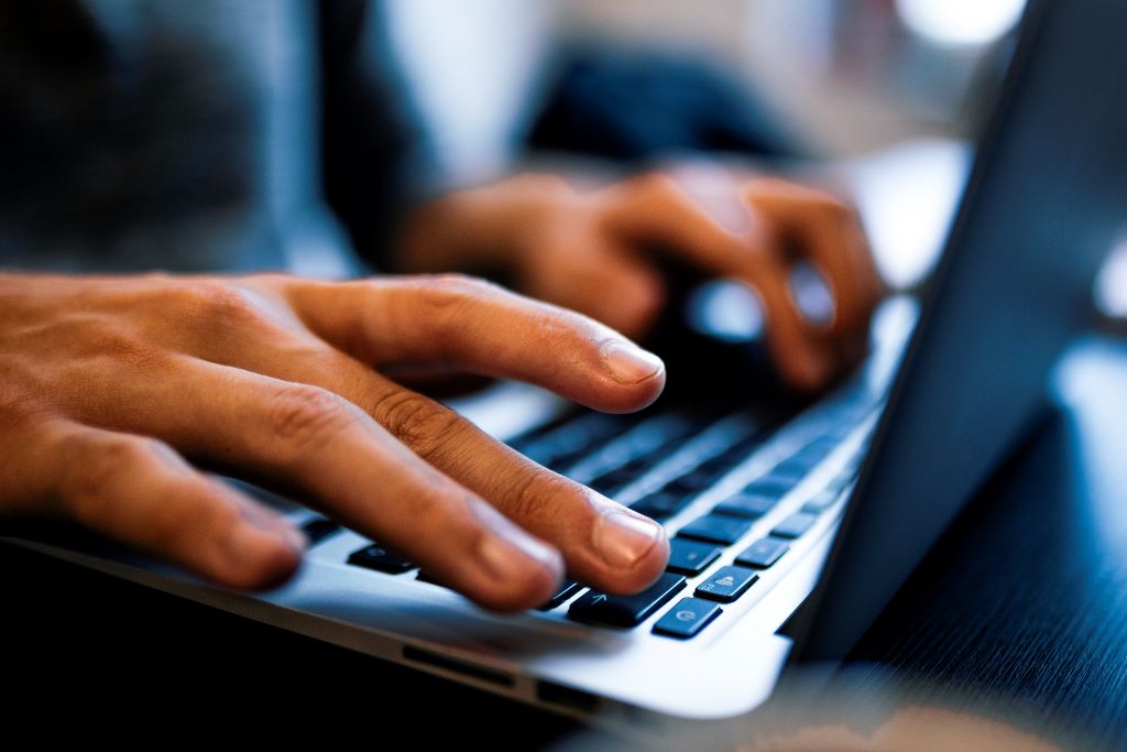 Close-up of an silver aluminium laptop rests on the lap of a student.  Two hands type on the keyboard.